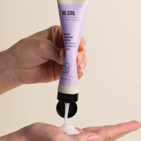 Hand holding a tube of RE:COIL skincare product with cream being dispensed onto another hand against a beige background.